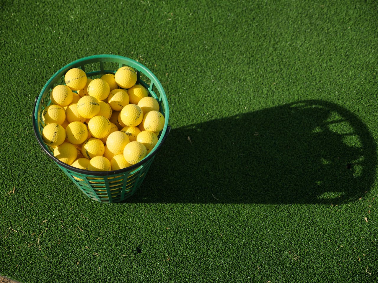 A basket full of yellow golf balls casting a shadow on vibrant green turf.